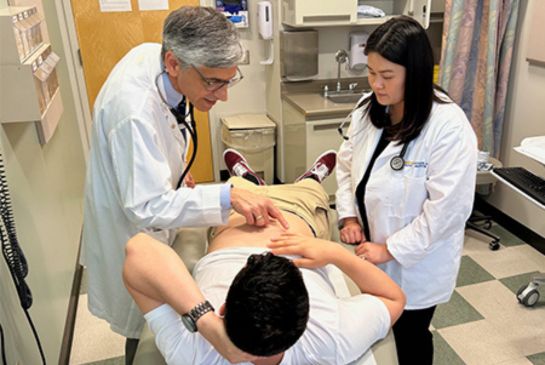 A doctor teaches a medical student how to examine a patient lying on an exam table in a clinic room during a training session.